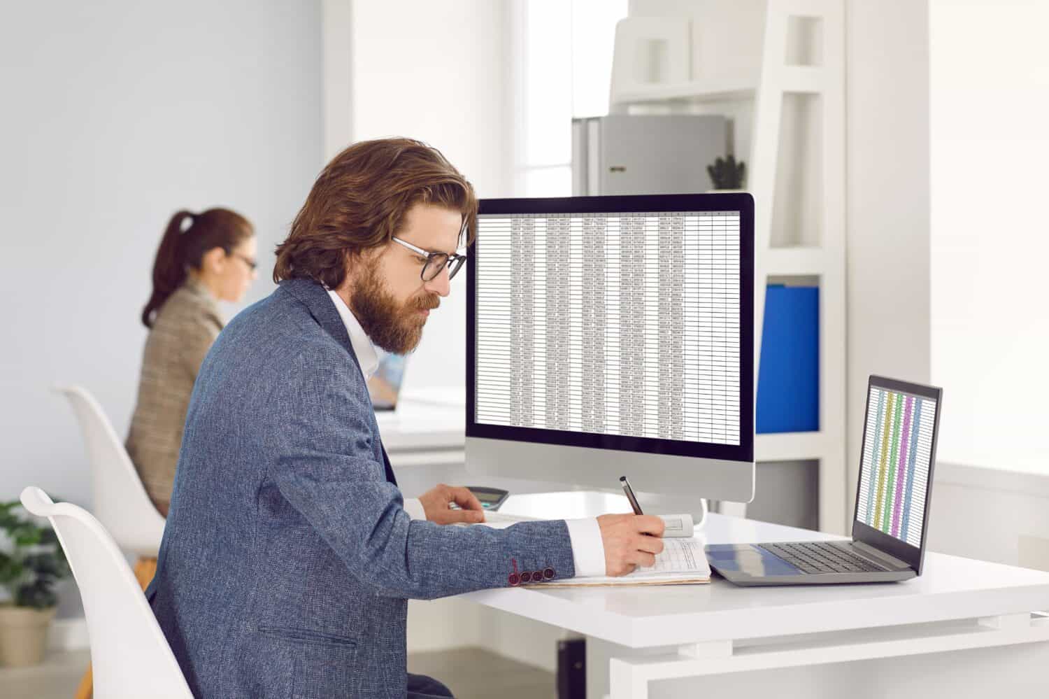 Adult Caucasian man works as accountant or auditor sits at table with monitor and laptop, fills out tables and makes entries in workbook calculating profits and taxes located in open space office