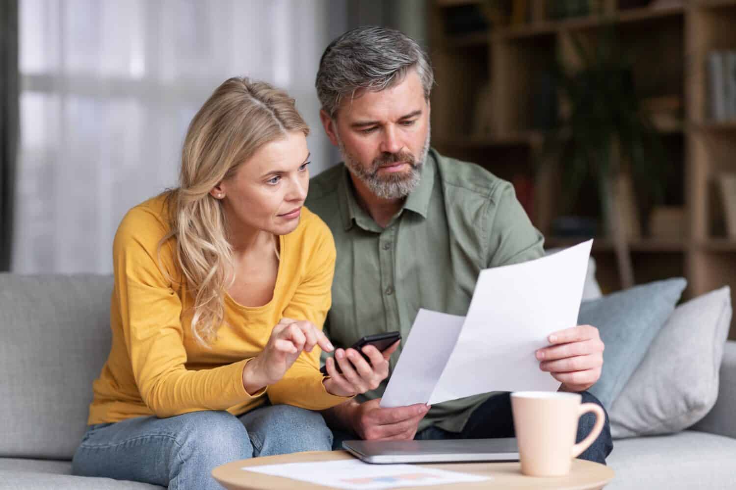 Married Middle Aged Couple Planning Budget Together, Reading Papers And Calculating Spends While Sitting On Couch In Living Room, Husband And Wife Checking Documents And Accounting Taxes, Closeup