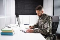 A man with short brown hair and a beard, wearing a green and brown camouflage military uniform, sits at a white desk. He is looking down and writing on a document held on a black clipboard with a silver pen. To his left is a stack of three books with light green, tan, and blue covers. A black computer monitor is visible behind the books. The background features light-colored walls and dark blinds.