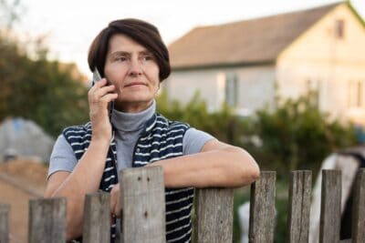 A middle-aged woman with short, dark brown hair and a pensive expression holds a smartphone to her ear, talking. She wears a gray turtleneck and a navy blue and white striped sleeveless jacket, leaning her left arm on a weathered wooden fence. In the soft-focus background, a light-colored house and green foliage are visible under an overcast sky.
