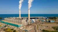 An aerial view of a large industrial power plant facility featuring two tall, light-colored smokestacks emitting white plumes of smoke. The facility has numerous grey and teal-colored buildings, connecting conveyor systems, and extensive paved areas. It is positioned next to a large blue body of water under a clear blue sky.