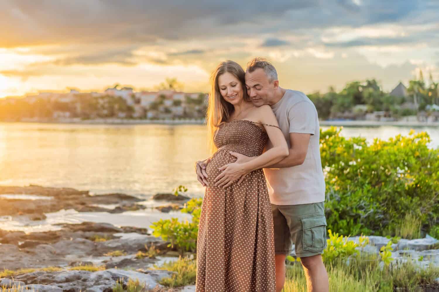 A happy, mature couple over 40, enjoying a leisurely walk on the waterfront On the Sunset, their joy evident as they embrace the journey of pregnancy later in life