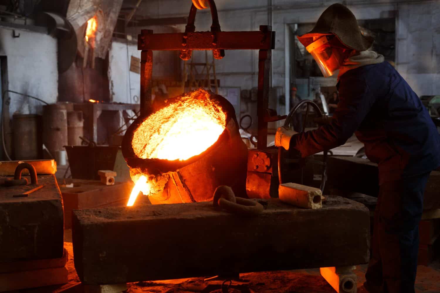 Industrial worker in protective gear pours molten metal at steel foundry. Manufacturing process in heavy industry with furnace. Skilled laborer handles high-temperature metallurgy, safety action.