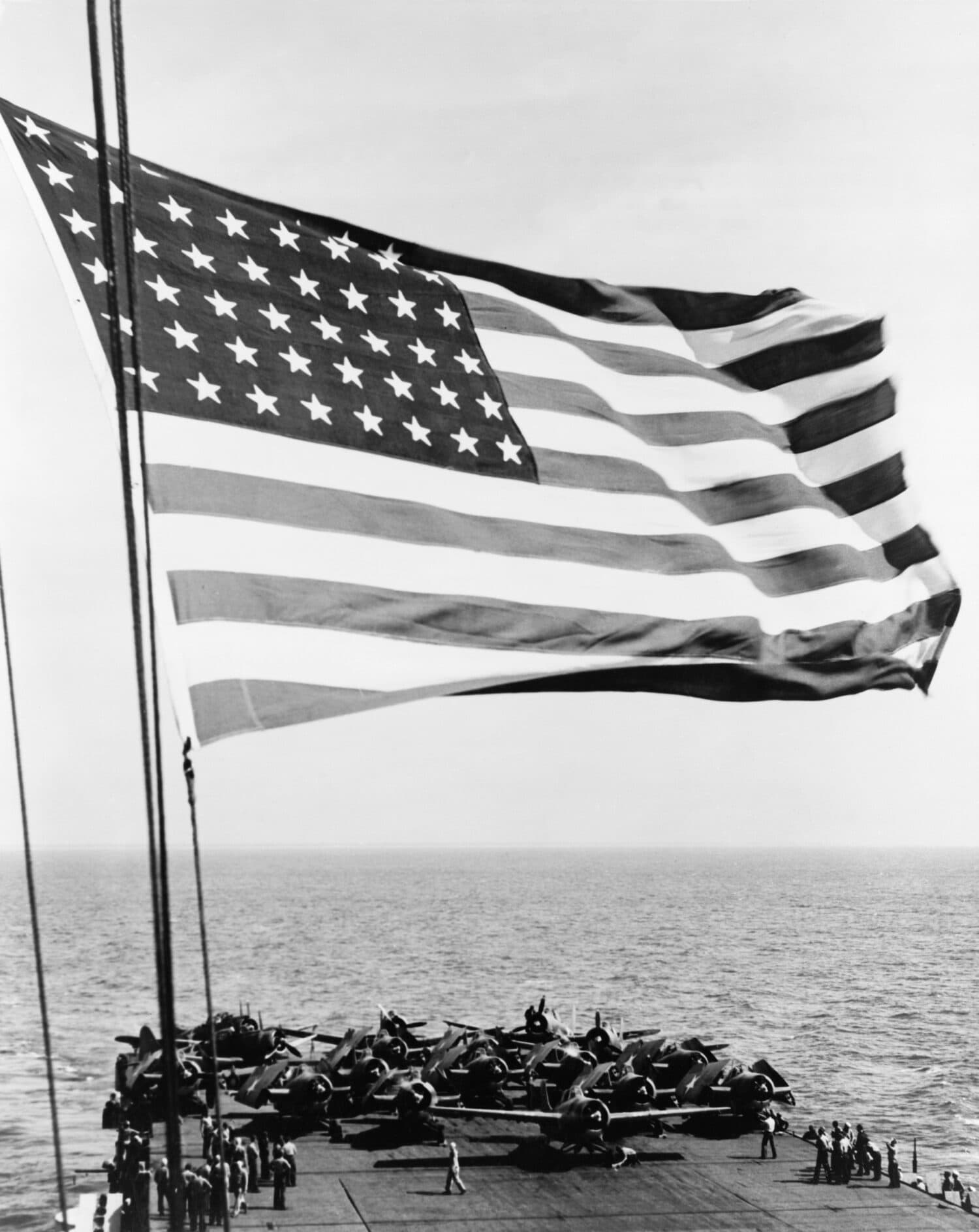 U.S. flag waving in foreground over flight deck of an escort carrier on July 16, 1943. Of the 151 aircraft carriers built in the United States during WWII, 122 were escort carriers.