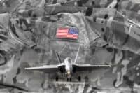A composite image featuring a desaturated military camouflage pattern in the background. In the upper-middle, a vibrant American flag patch is sewn onto a military uniform, showing its red and blue colors prominently. Overlaid on the lower half is a gray fighter jet, likely an F/A-18, facing directly forward with landing gear extended and bright landing lights illuminated, appearing ready for action.