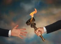 A close-up shot of two hands against a blurred, gradient background of blues, grays, and browns. On the right, a person's arm, dressed in a dark suit jacket with a white shirt cuff, holds a golden, antique-style torch with vibrant orange flames emanating from its bowl. On the left, another person's bare hand, also wearing a dark suit jacket and white cuff, is outstretched with an open palm, appearing to accept the torch.