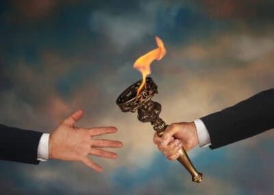 A close-up shot of two hands against a blurred, gradient background of blues, grays, and browns. On the right, a person's arm, dressed in a dark suit jacket with a white shirt cuff, holds a golden, antique-style torch with vibrant orange flames emanating from its bowl. On the left, another person's bare hand, also wearing a dark suit jacket and white cuff, is outstretched with an open palm, appearing to accept the torch.