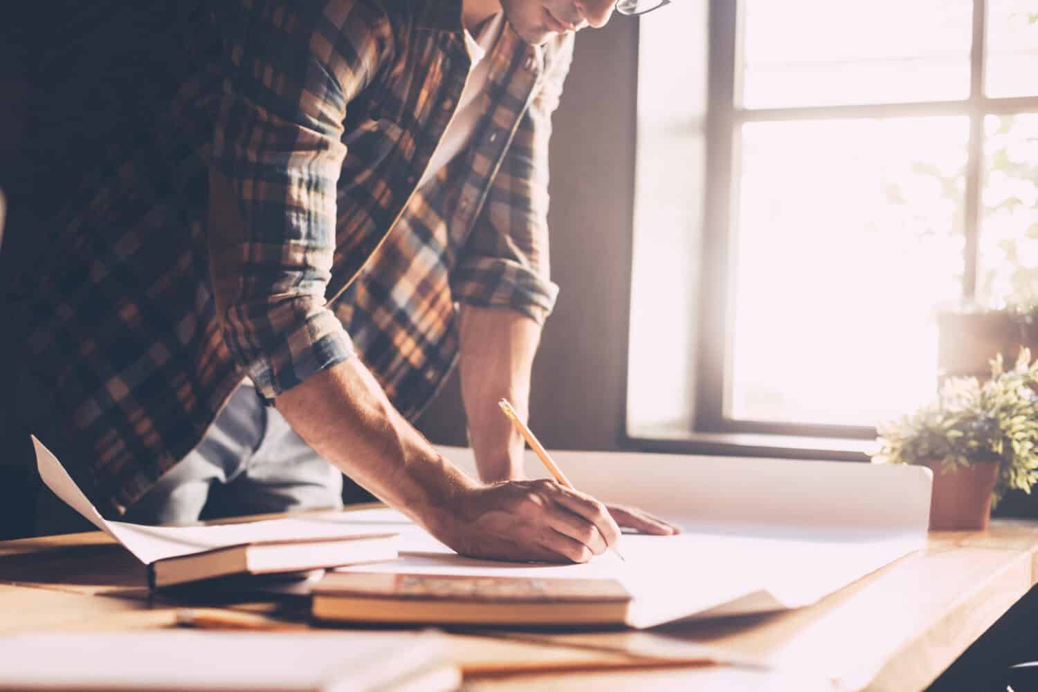 Working with passion. Close-up of confident young man in casual wear sketching on blueprint while standing near wooden desk in creative office