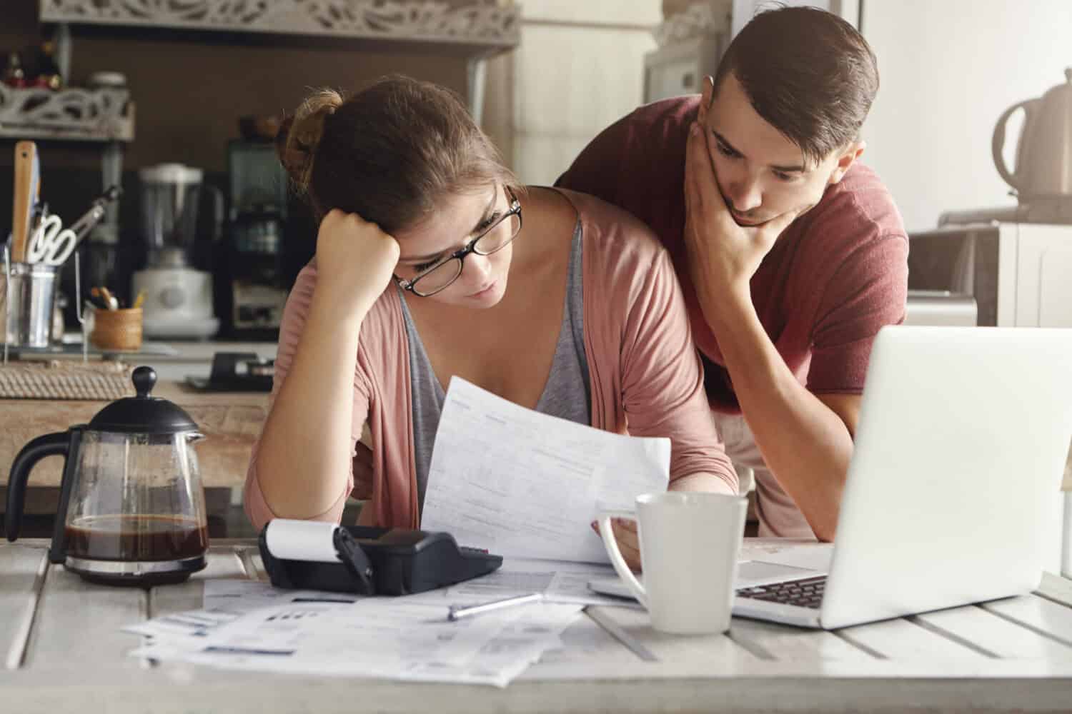 Young stressed Caucasian couple facing financials troubles, sitting at kitchen table with papers, calculator and laptop computer and reading document from bank, looking frustrated and unhappy