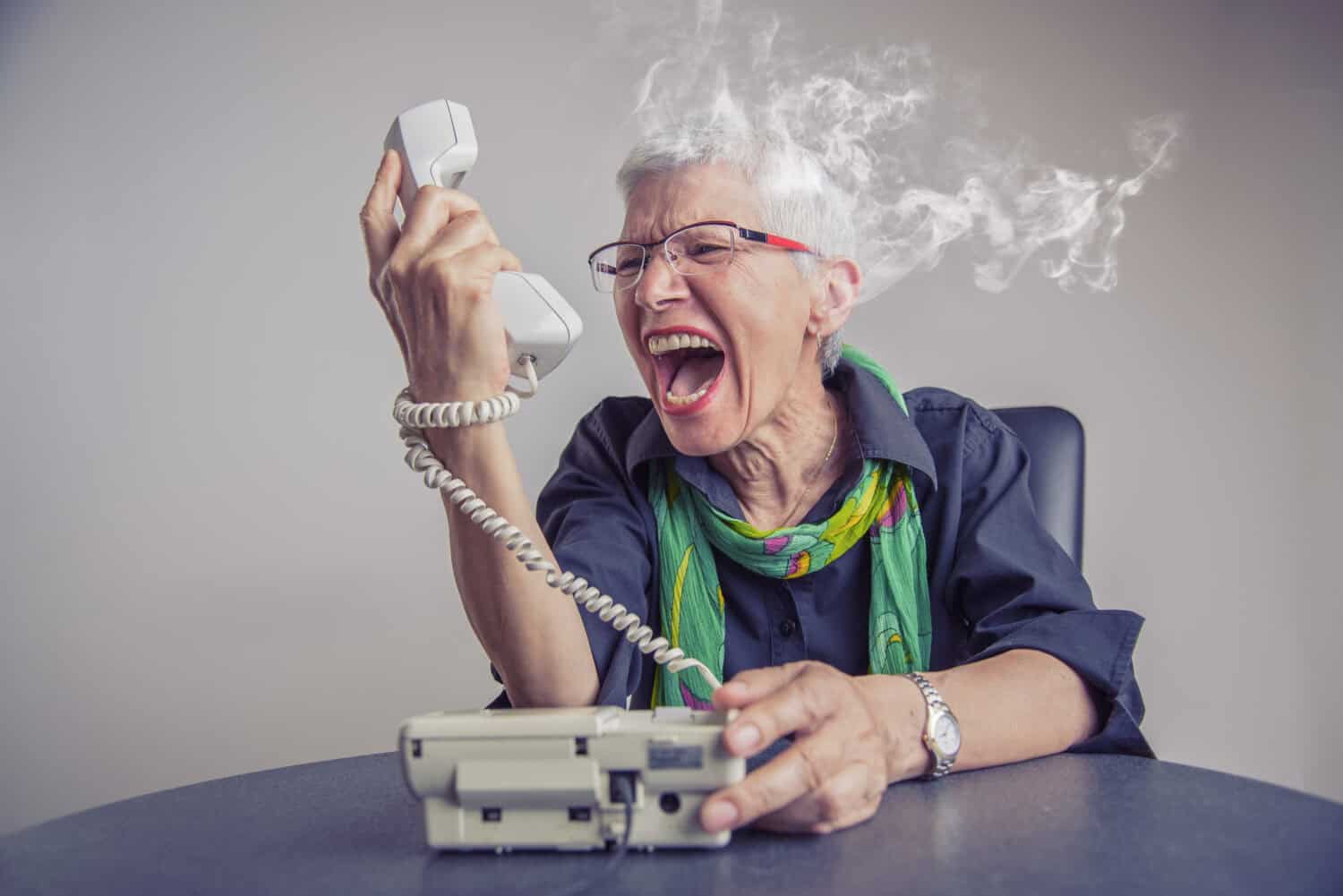 Angry, enraged senior woman yelling at a landline office phone, unhappy with customer service provided by the agent on the other side, giving off steam and smoke