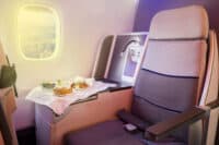 An interior view of a luxurious airplane cabin features a comfortable, grey and blue-striped seat in the foreground. A tray table extends, holding a served meal with bread rolls, a fresh salad, and a main dish on a white cloth. Through the oval window, bright sunlight illuminates a vast expanse of white clouds. Next to the seat, a panel with power outlets and an adjustable reading light is visible, all under soft purple cabin lighting.