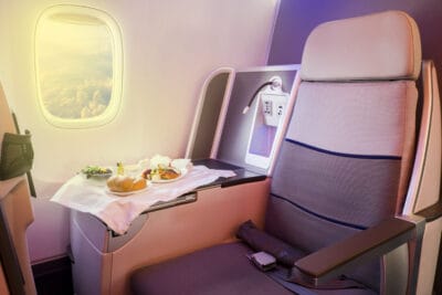 An interior view of a luxurious airplane cabin features a comfortable, grey and blue-striped seat in the foreground. A tray table extends, holding a served meal with bread rolls, a fresh salad, and a main dish on a white cloth. Through the oval window, bright sunlight illuminates a vast expanse of white clouds. Next to the seat, a panel with power outlets and an adjustable reading light is visible, all under soft purple cabin lighting.