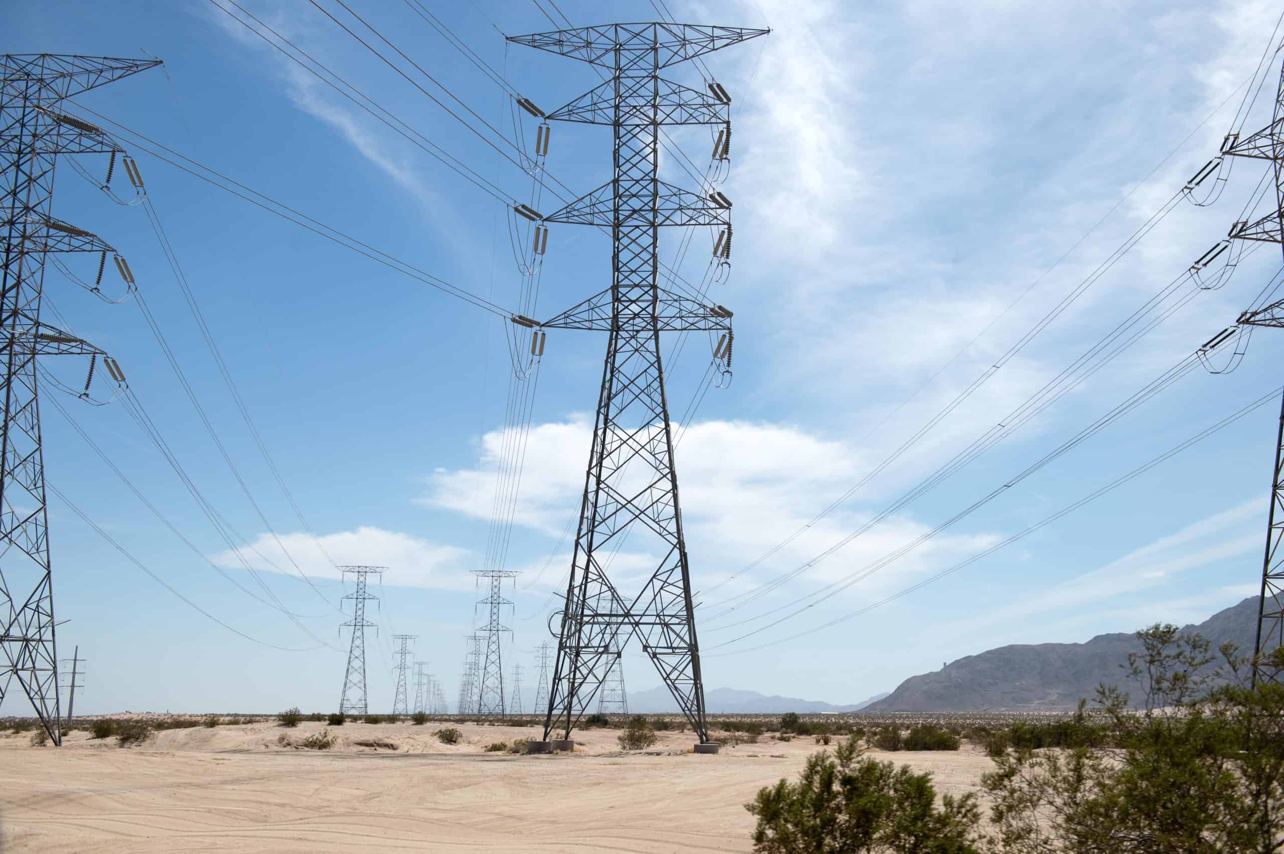 A panoramic view of multiple towering metal electrical transmission pylons with power lines stretching across a wide, flat desert landscape under a clear blue sky. Sparse green bushes dot the foreground, and mountains are visible in the far background.