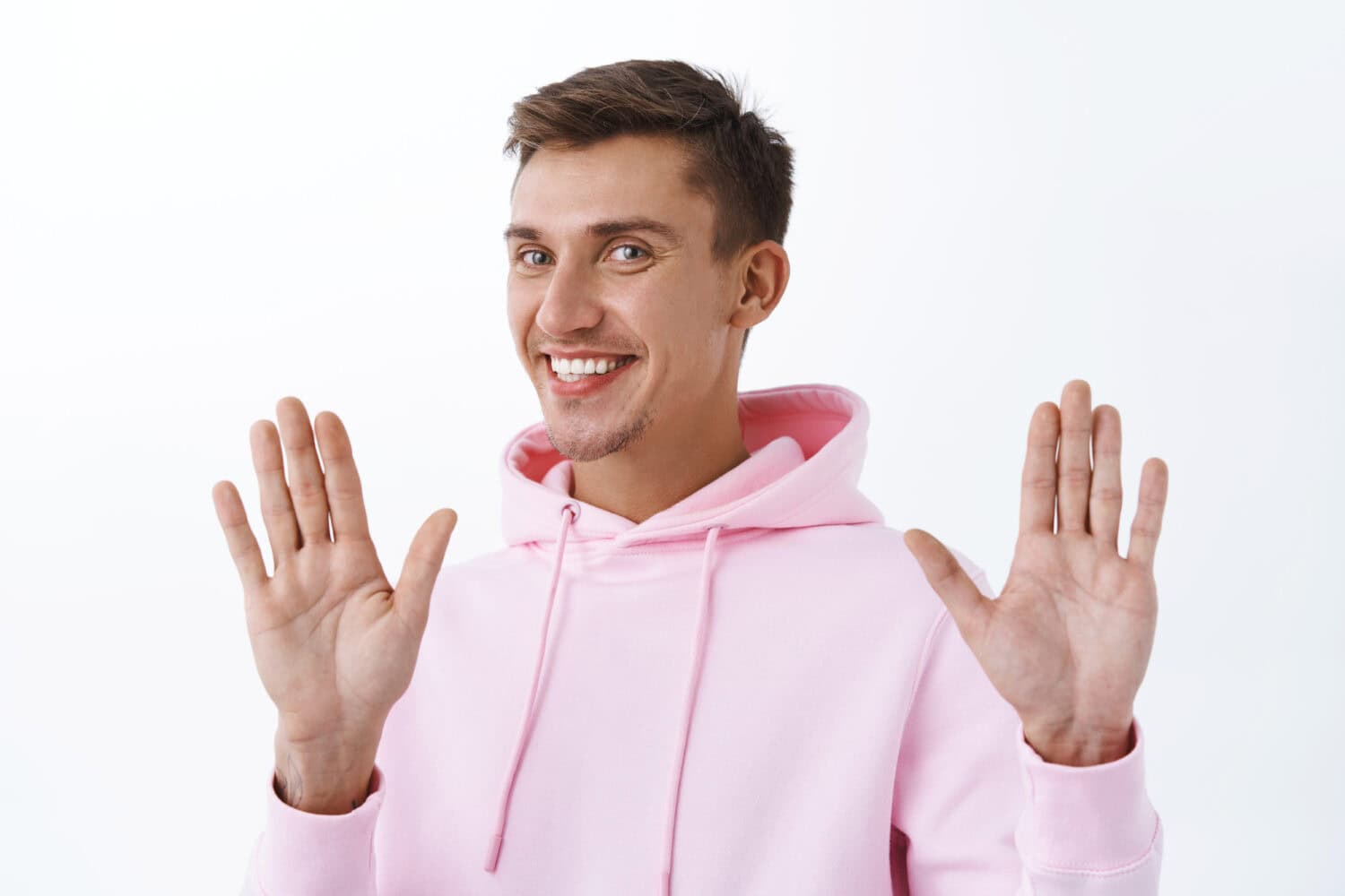 No thank you, I am okay. Portrait of handsome smiling blond man in pink hoodie, raising hands in surrender or sorry gesture, refuse or rejecting offer politely, standing white background