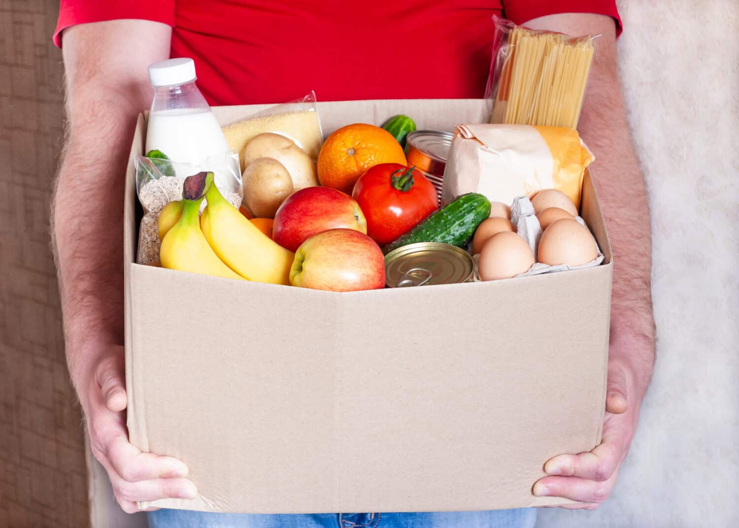 Grocery delivery courier man in red uniform holds cardboard box with fresh vegetables, fruits and other food. Express food delivery, donation concept.
