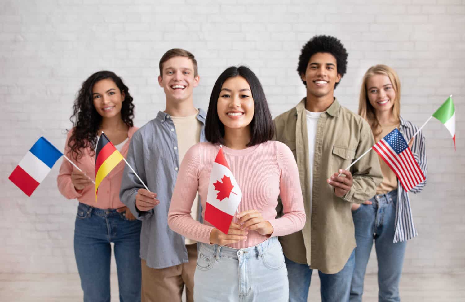 Young students of language school celebrate holiday. Happy millennial peoples of different nationalities have fun on study and hold little flags on brick white wall background, studio shot, free space