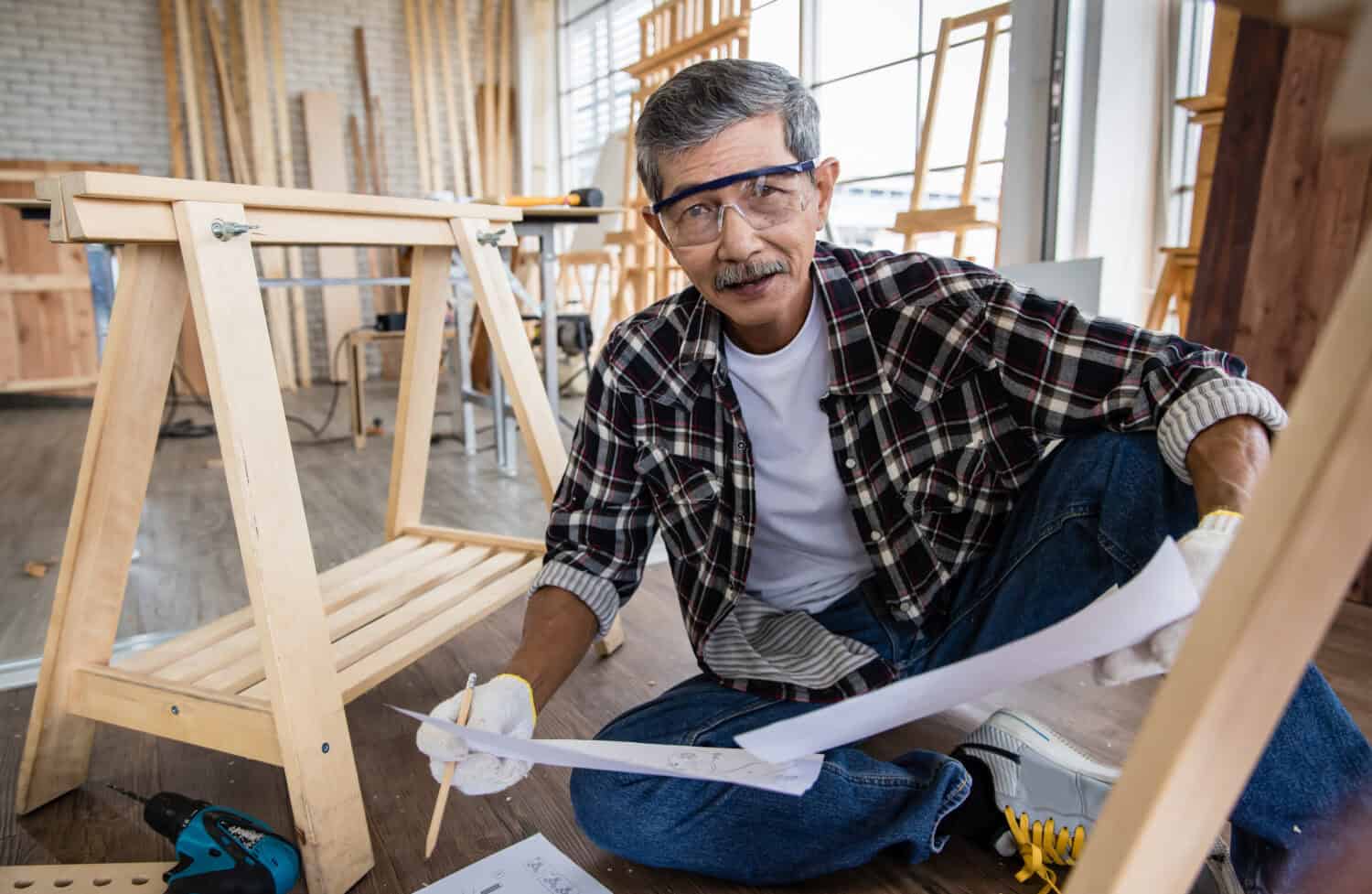 Aged Asian man with blueprint reading data on laptop while making timber furniture in joinery. Older retired people hobby and DIY in free time concept.