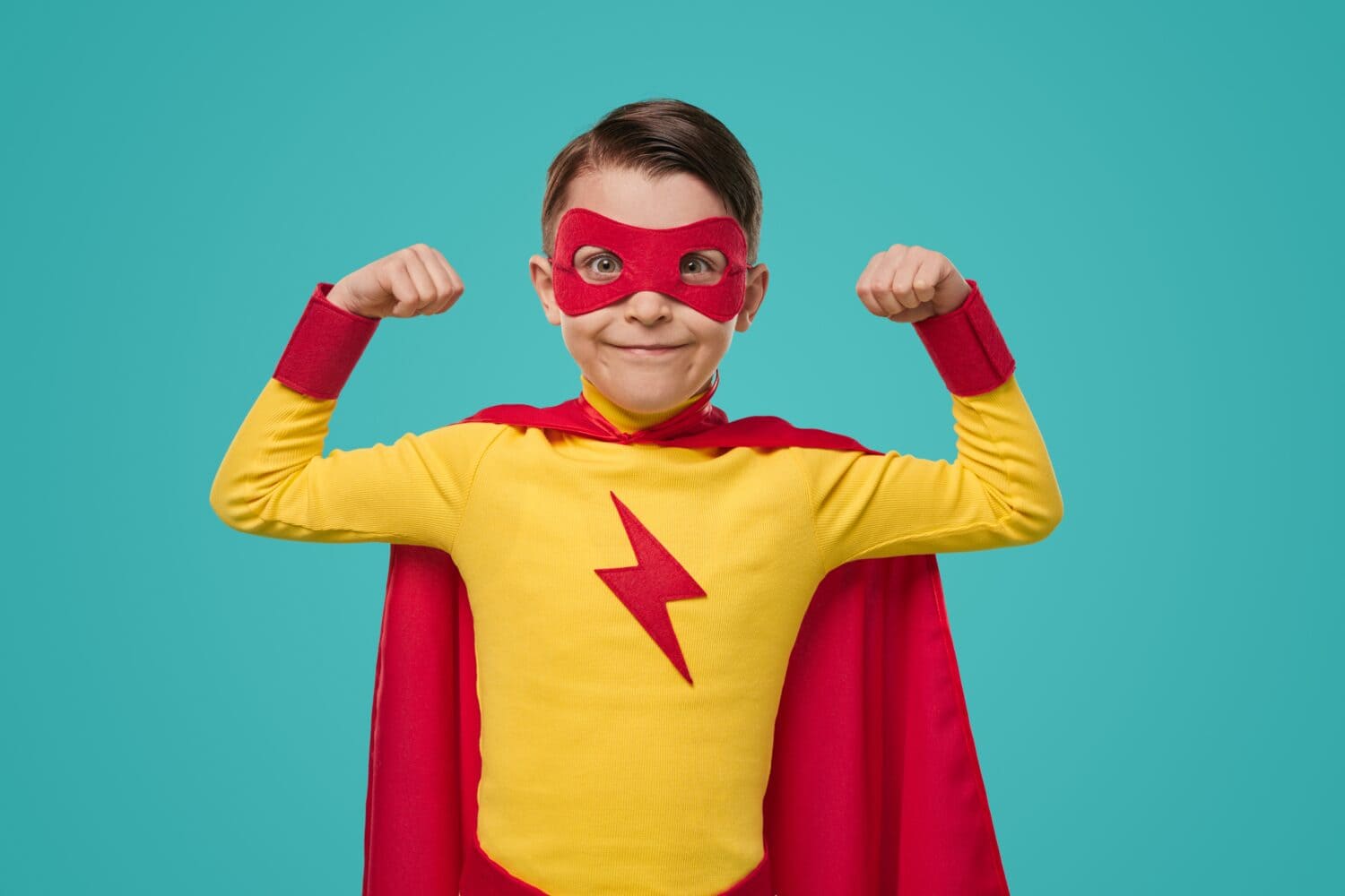 Content superhero kid in red and yellow cape and mask demonstrating power while looking at camera against blue background in studio