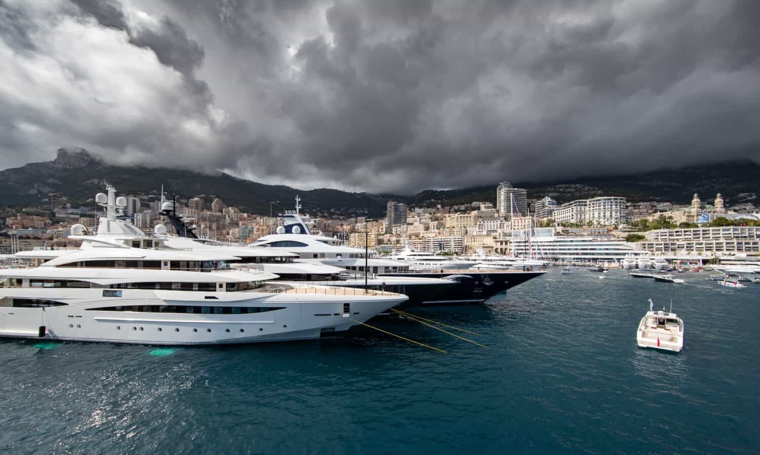 A lot of huge yachts are in port of Monaco at storm weather, mountain is on background, glossy board of the motor boat, megayachts are moored in marina, sun reflection on glossy board