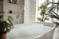 Soft native hues organic shapes look of bathroom with big window oval bathtub in neutrals tones. Green palm plants candles bubblebath leasure and relaxation skin selfcare wellness luxury living