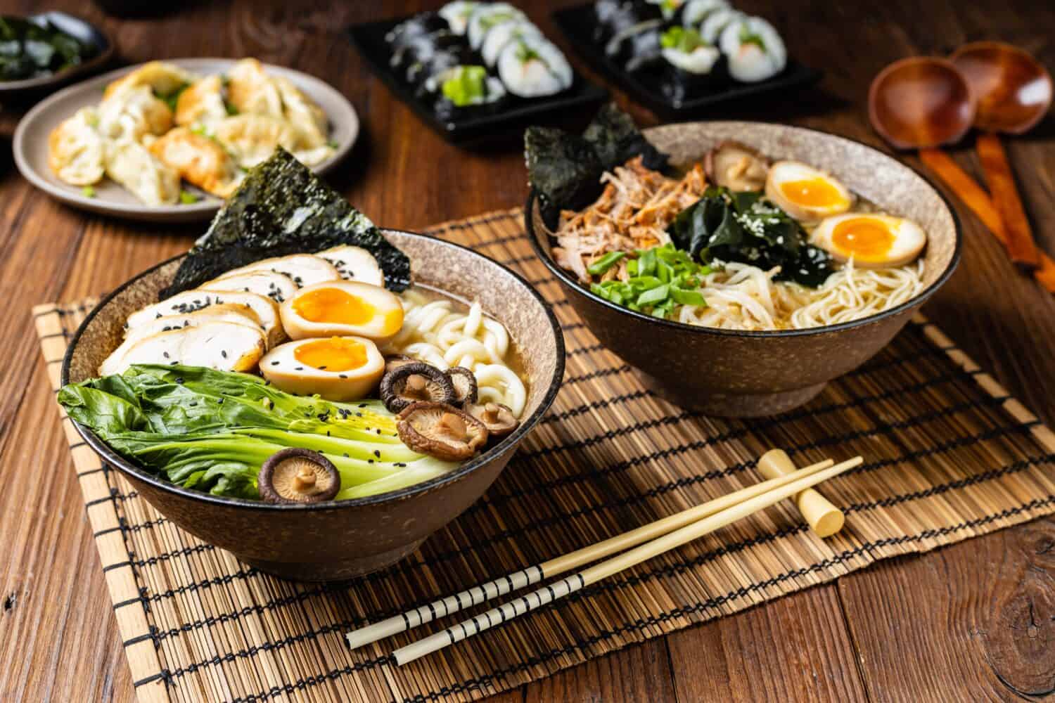 Traditional ramen with jerked pork or chicken. With udon or ramen noodles. Served in classic bowls. Gyoza dumplings and mushrooms in the background. Natural wooden background.