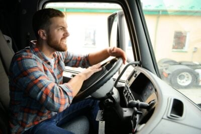 Side profile of a bearded man in a blue and orange plaid shirt, sitting in the driver's seat of a truck. His face shows an expression of intense anger or stress, with gritted teeth and a furrowed brow, as his hands grip the steering wheel. The exterior visible through the window is blurred.