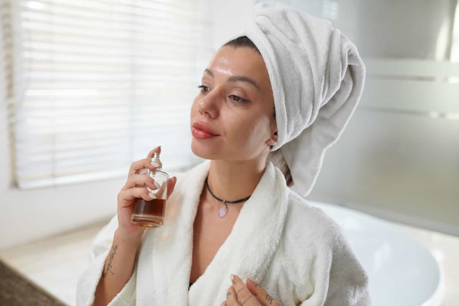 Young pleased woman in bathrobe spraying new fragrance from bottle on her skin and enjoying smell of perfume while standing in bathroom