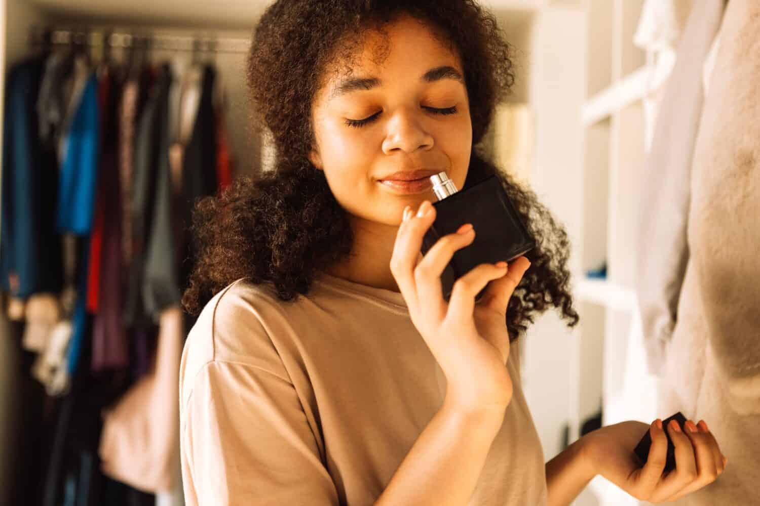 Close-up of charming afro american teenager holding glass bottle and sniffing perfume Attractive African girl in casual clothes has closed her eyes and is enjoying the scent of perfume Modern interior