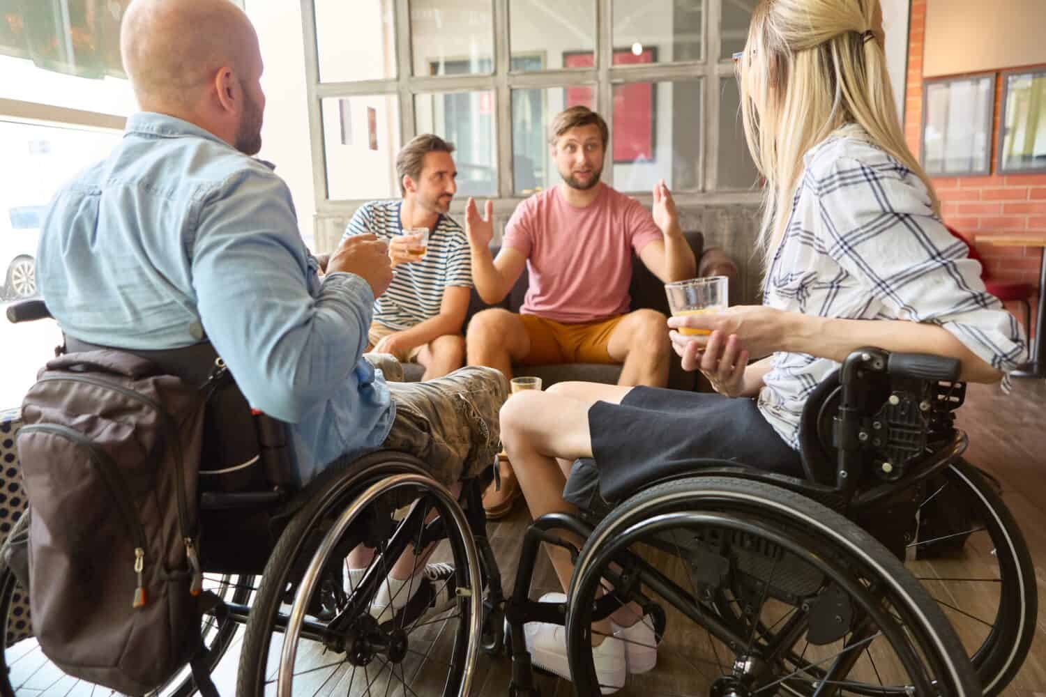 A group of friends, including people who use wheelchairs, engaged in lively conversation and enjoying drinks together in a sunny café setting.