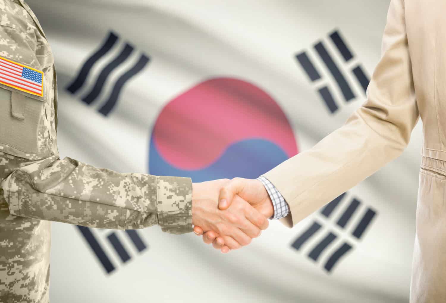 American soldier in uniform and civil man in suit shaking hands with adequate national flag on background - South Korea