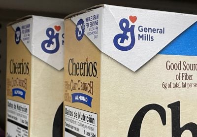 Several brown and white boxes of General Mills Cheerios Oat Crunch Almond cereal are stacked on a store shelf. The blue General Mills logo, with a stylized 'G' and a red heart, is prominently displayed on the upper part of the boxes. Text visible includes 'WHOLE GRAIN PER SERVING' and 'Datos de Nutrición' in Spanish.
