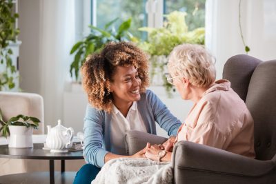 A smiling younger woman with curly brown and blonde hair, wearing a white shirt and blue cardigan, holds the hand of an older woman with short blonde hair and glasses, wearing a light pink shirt. They are sitting in comfortable armchairs, facing each other in a bright room with houseplants and a round side table with a teapot.