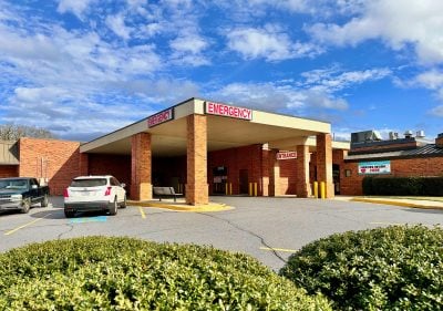 A wide exterior shot of a brick hospital building's emergency room entrance on a sunny day with a blue sky and scattered white clouds. A covered drive-through area is supported by four brick columns, with red 'EMERGENCY' signs overhead and an 'ENTRANCE' sign near the main doors. A banner on the brick wall to the right reads 'HEROES WORK HERE' with a heart and heartbeat icon. A dark green pickup truck is parked on the left, and a white SUV is on the right, with green bushes in the foreground.