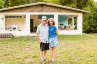 A smiling older couple stands arm-in-arm in a verdant green lawn in front of a simple, single-story white house with an open porch. The man, wearing a light-colored fedora, light blue collared shirt, dark shorts, and sandals, has his arm around the woman, who wears a blue t-shirt, a white and blue floral skirt, and sandals. The house has a light brown thatched-style roof and open windows revealing a washing machine on the left and a sitting area with light blue walls on the right. Lush green trees and foliage are visible in the background under a bright sky.