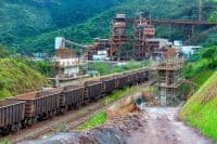 A panoramic view of a large iron ore mining complex. In the foreground, a long train with multiple dark-colored open-top freight cars sits on a railway track. To the left, lush green hills rise steeply. In the midground and background, an extensive industrial facility with tall, rusty-brown metal structures, conveyor belts, and processing units is visible, also surrounded by green, forested mountains. A construction site with scaffolding is present near the railway tracks on the right.