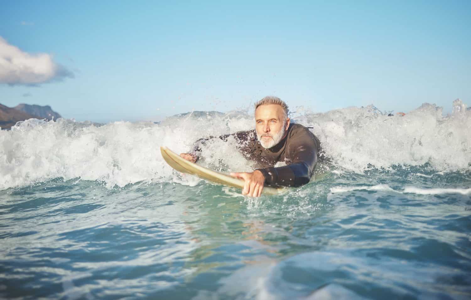 Ocean waves, senior man surfing on the beach and healthy fitness lifestyle on summer vacation in Australia. Senior surfer swimming with surfboard, exercising with sea water and relaxing on retirement travel vacation