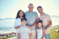 A smiling multi-generational family of five stands outdoors against a bright blue sky and ocean background. The group consists of an older man with glasses in the center, flanked by a younger woman with curly hair and a younger man. Two children, a boy and a girl, stand in front. The family is looking directly at the viewer and smiling, with a rocky shoreline and distant mountains visible behind them.
