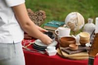 A person in a white shirt and light-colored pants places a small dark bowl onto a stack of plates on a red table. The table is laden with various household items including white and wooden mugs, bowls, stacks of books, a vintage globe, and silverware. In the background, green grass is visible, and a decorative heart made of wood pieces sits on the ground. The setting suggests an outdoor market or yard sale.