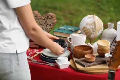 A person in a white shirt and light-colored pants places a small dark bowl onto a stack of plates on a red table. The table is laden with various household items including white and wooden mugs, bowls, stacks of books, a vintage globe, and silverware. In the background, green grass is visible, and a decorative heart made of wood pieces sits on the ground. The setting suggests an outdoor market or yard sale.