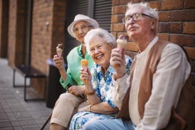 Three older adults, two women and one man, sit smiling outdoors on a paved area next to a brick wall, each holding an ice cream cone. The woman in the center with white hair is laughing. She wears a blue and white patterned shirt. To her left, an older woman in a green top and white hat holds a chocolate-dipped cone. To her right, an older man with glasses and a mustache holds a vanilla and chocolate swirled cone. They appear relaxed and happy.