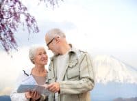 An elderly couple, a woman with white hair smiling up at a man with glasses and a beard, jointly look at a folded map. The woman holds a 'City Guide' booklet. In the background, a blooming tree with purple flowers frames the top left, and a snow-capped mountain is visible under a cloudy sky.