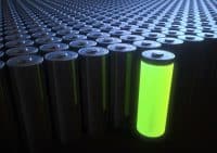 A large grid of cylindrical batteries, mostly dark grey or black, with metallic tops. One battery in the front right glows vibrantly with a bright neon green light from within, illuminating its surroundings slightly. The rows of batteries recede into the dark background, creating a sense of depth and multitude.