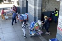 An overhead view of an outdoor area outside a warehouse store shows several people. On the left, an older woman in a maroon jacket and an older man with glasses are near a shopping cart filled with groceries. In the center, a man in a blue shirt and gloves pushes an orange pallet jack stacked with large packages of bath tissue and bottled water. To his right, a person in a plaid shirt and gloves pushes an overflowing shopping cart with bath tissue, drinks, and a cake. Further right, a woman in a straw hat pushes a shopping cart towards an open bay door, while a worker in a yellow high-visibility vest stands by a red floor machine inside the building entrance. The ground is grey concrete, and the building exterior is grey block.