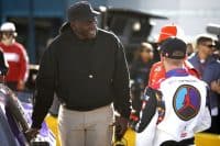 Michael Jordan, a Black man, is smiling and wearing a black hoodie with an Air Jordan logo and a black baseball cap. He is looking towards and engaging in conversation with a race car driver whose back is to the camera. The driver's white, red, and blue racing suit prominently displays 'TYLER REDDIC' and a large circular Jordan Brand logo featuring a red silhouette on a light blue and purple background. A person in an orange 'DOORDASH' cap is partially visible in the background, to the right.