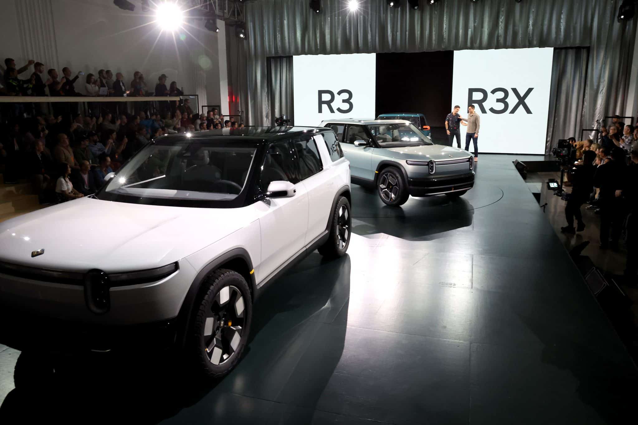 A white Rivian R3 electric vehicle with a black roof is in the foreground, facing left. Behind it and slightly to the right is a lighter grey Rivian R3X electric vehicle. Both cars are on a dark stage, illuminated by bright spotlights. In the background, large white screens display the text 'R3' and 'R3X' in bold black letters. An audience of people is visible in tiered seating and on the ground, watching the presentation.