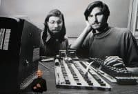 A smiling Steve Jobs, wearing a black turtleneck and jeans, stands on a stage, gesturing with his hands. He is superimposed over a large, grainy black and white photograph. This background image depicts a younger Steve Jobs with a mustache, resting his head on his hand while looking forward, alongside a younger Steve Wozniak with a beard and glasses, looking down at an early computer circuit board. To the far left of the background image, an old computer monitor displays the repeated text 'BYTE SHOP'.