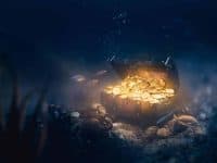 An open, ornate wooden treasure chest sits on the seabed, overflowing with shiny gold coins that emit a bright, warm light. Bubbles rise from the chest into the dark blue water where several small fish swim, with various corals and rocks littering the ocean floor.
