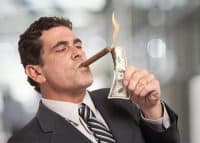 A man in a dark suit and striped tie is shown in profile, lighting a cigar held in his mouth with a burning US $100 bill. Flames rise from the top of the bill as he holds it up to the cigar. The background is a soft, out-of-focus grey.