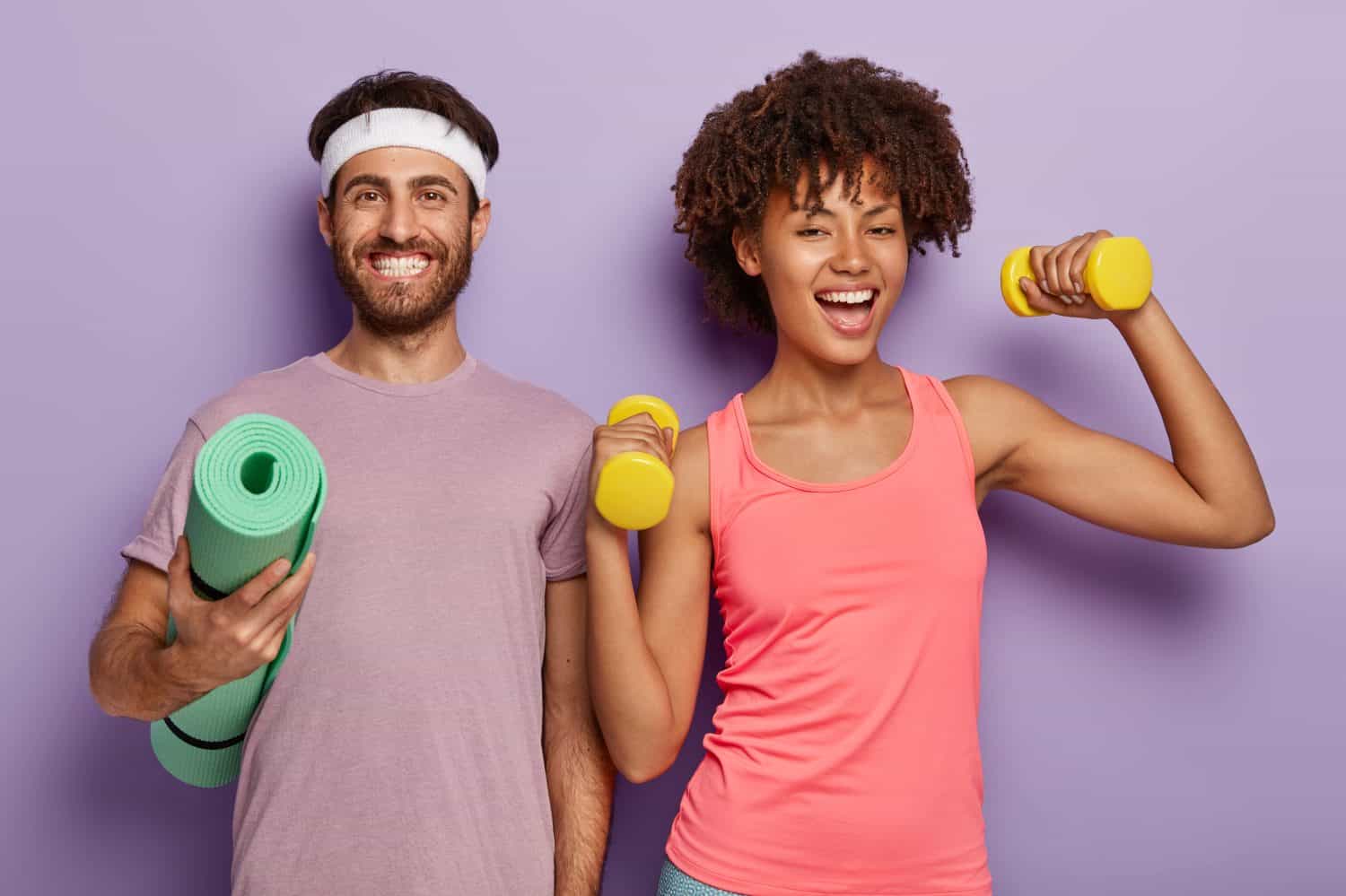 Improve your muscles. Sporty woman training with dumbbells, looking cheerful, her husband standing next to her, holding the rolled up fitness mat, enjoying athletic training together, isolated on purple background