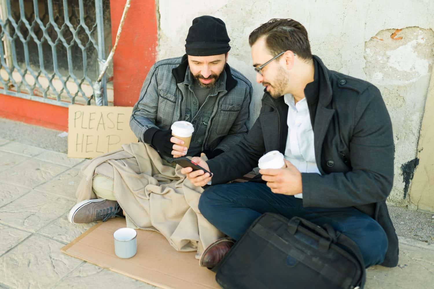 Smiling showing pictures or photos in smartphone to homeless man with beard while drinking coffee together